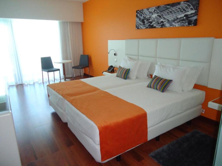 Quarto de hotel com duas camas, tapa pés laranja, almofadas decorativas e cabeceira de cama estofada branca