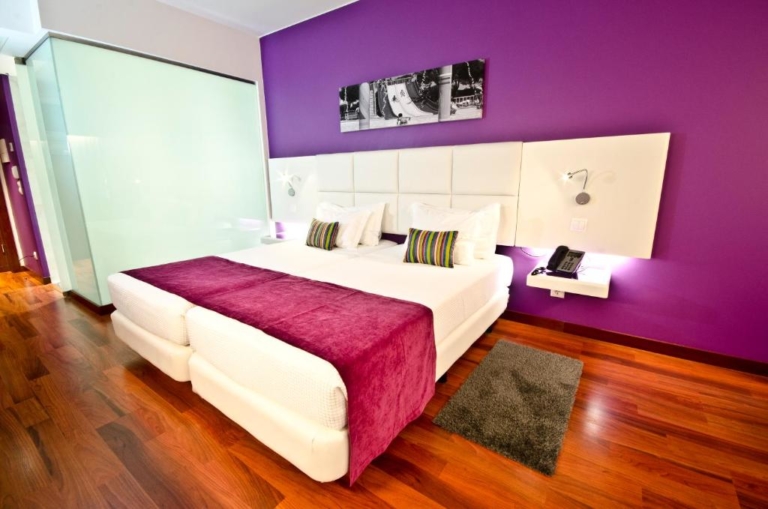 Quarto de hotel com duas camas, tapa pés rosa e almofadas decorativas, cabeceira de cama estofada em branco