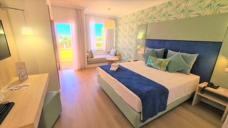 Quarto de hotel com cabeceira de cama verde água e azul, cama com almofadas decorativas azuis e com padrões em tons de verde