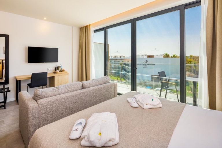 Quarto de hotel com cabeceira de cama, base, colchão tapa pés creme e almofadas decorativas e cortinado em ondas