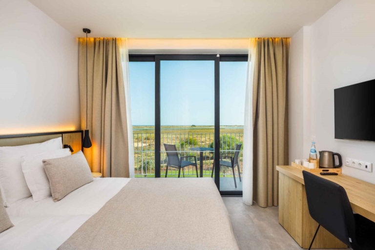 Quarto de hotel com cabeceira de cama, base, colchão tapa pés creme e almofadas decorativas e cortinado em ondas