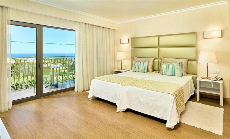 Quarto de hotel com cabeceira de cama verde, duas camas com cobre pés, almofadas decorativas lisas e com padrão em tons de creme e verde