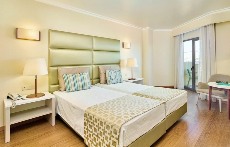 Quarto de hotel com cabeceira de cama verde, duas camas com cobre pés, almofadas decorativas lisas e com padrão em tons de creme e verde