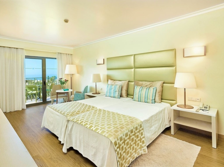 Quarto de hotel com cabeceira de cama verde, duas camas com cobre pés, almofadas decorativas lisas e com padrão em tons de creme e verde